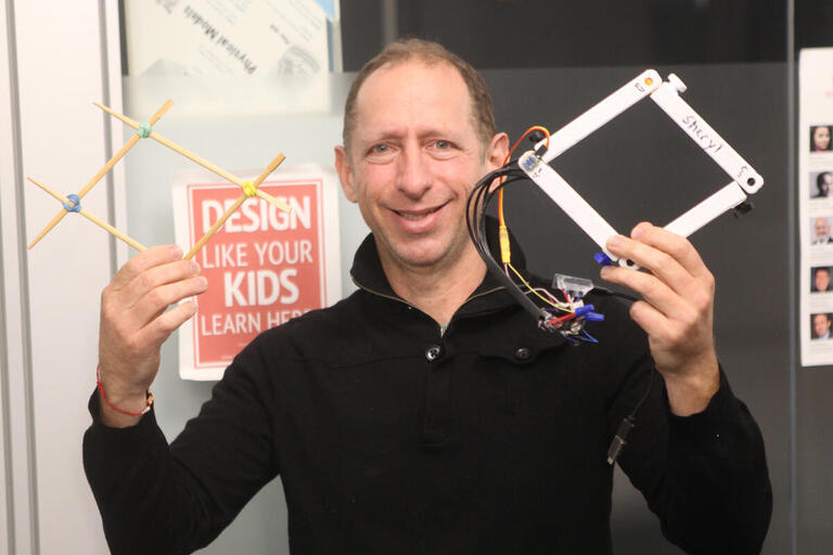 Professor Dor Abrahamson is holding a model quadrilateral made from wooden chopsticks and rubberbands, and a 3D printed version that plugs into a computer.