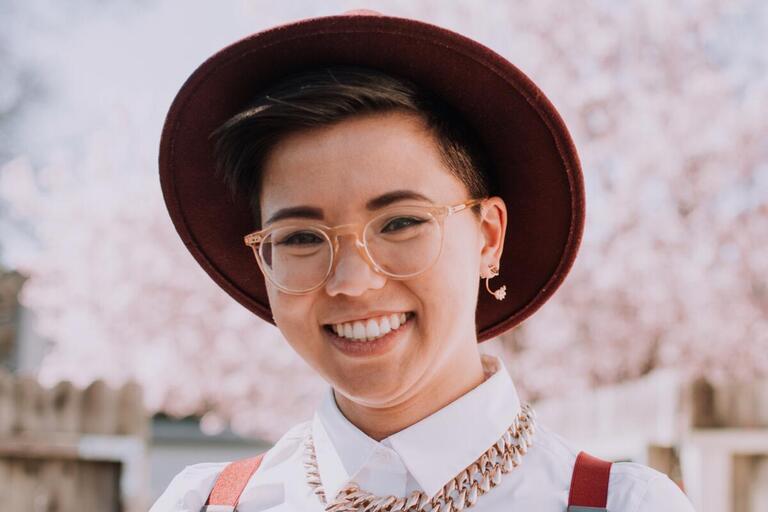 Mers Tran smiling at camera wearing bolero hat, glasses, white button up top, gold choker, red suspenders