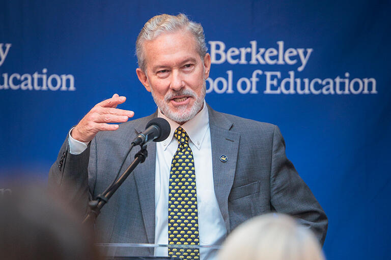 chancellor rich lyons at the podium speaking to the audience with right hand raised in a gesturing movement standing in front of a back drop with the  u c berkeley school of education logo