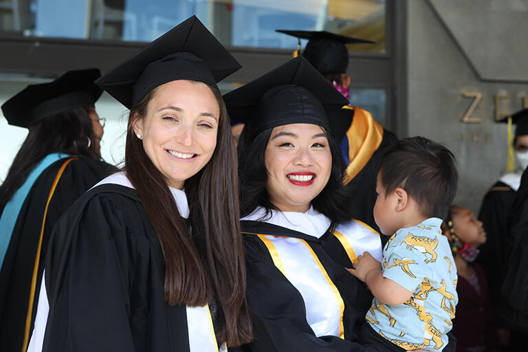 two masters degree candidates smiling and one is holding a toddler 