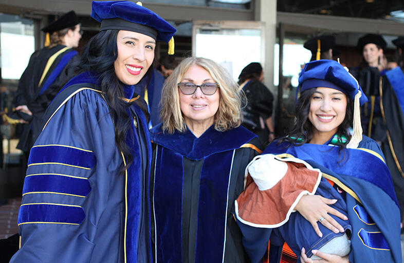 Assistant dean Kris Gutierrez standing in the middle of two students who earned their doctoral degree. student right is holding an infant