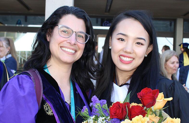 a graduate holding flowers standing next to a faculty member and both are smiling at the camera