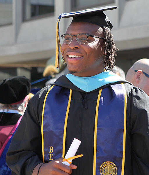 a master's degree student wearing regalia smiling and holding a paper scroll representing their diploma