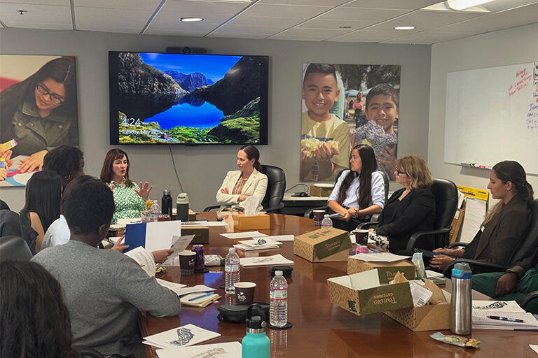 eight people sitting around a table looking at the person speaking at the head of the table with lunch boxes, drinks and papers in front of them
