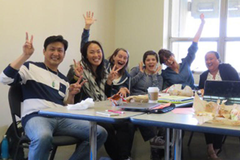 six students sitting at table smiling at camera and some holding up two fingers as peace sign