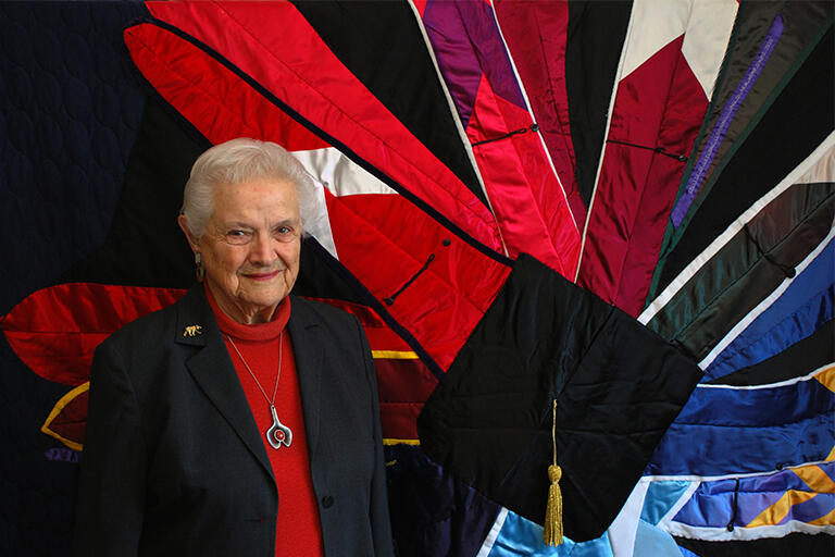 Pat Cross standing in front of a quilt made from many of her college regalia hoods