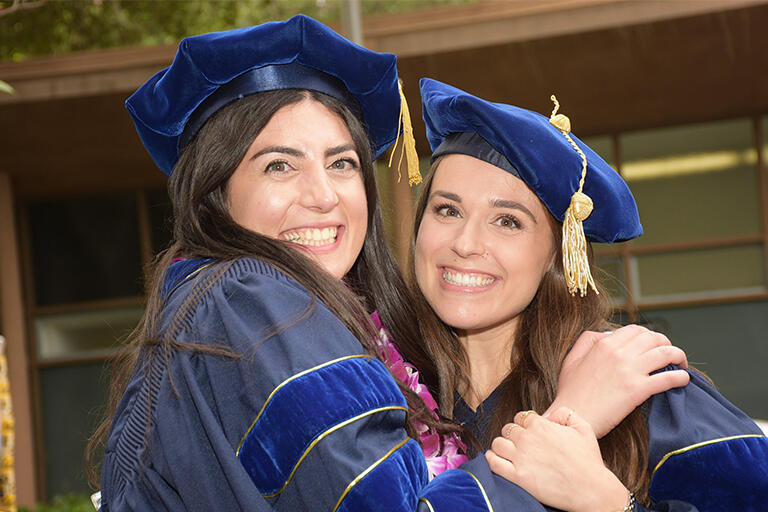 two doctoral graduates wearing their blue regalia hugging and smiling at camera