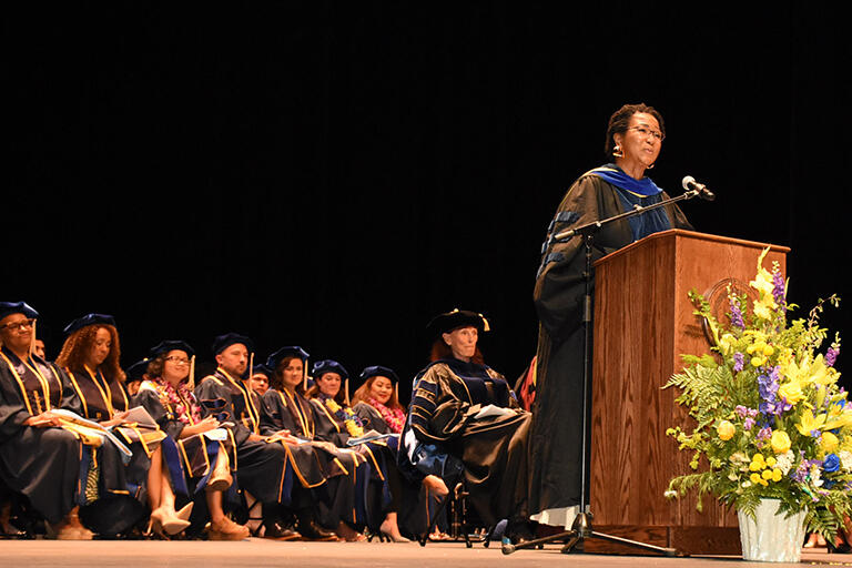 doctor mae jemison standing at podium with some faculty and graduates sitting on stage behind her