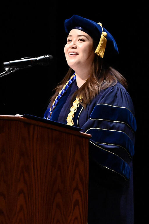 meg stomski wearing doctorate regalia at podium delivering a speech