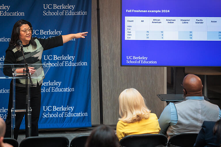 Rebecca Cheung at podium speaking to the audience and pointing to a slide on the right that has some admissions data