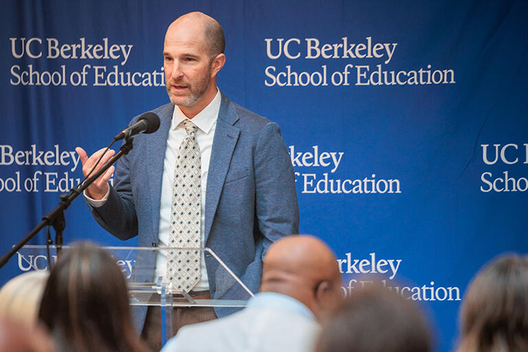 joshua daniels at the podium speaking to the audience standing in front of a back drop with the u c berkeley school of education logo