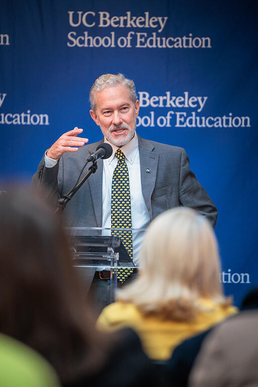 chancellor rich lyons at the podium speaking to the audience with right hand raised in a gesturing movement standing in front of a back drop with the  u c berkeley school of education logo