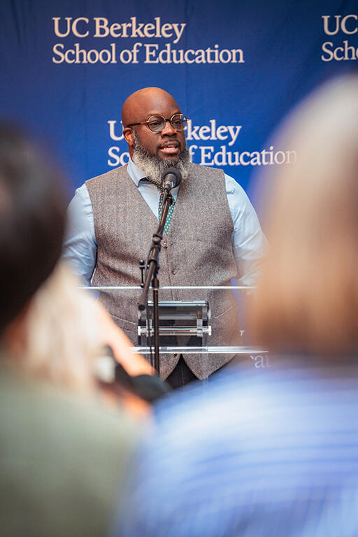 femi ogundele at the podium speaking to the audience standing in front of a back drop with the  u c berkeley school of education logo