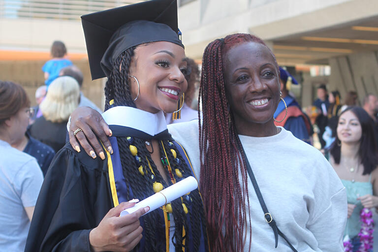 a person on the right with their arm around a graduate who is holding a paper scroll after the ceremony and both are looking to the right at someone taking their picture