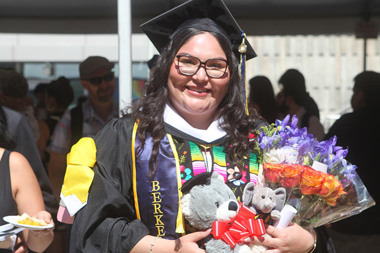 a graduate wearing their regalia and smiling at the camera holding flowers and two small stuffed animals