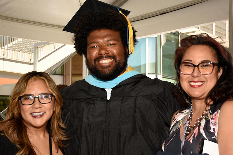 a master's degree student wearing their regalia and two people on either side of them with everyone smiling at camera