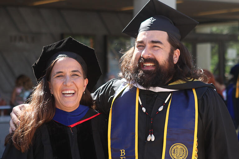 a graduate on the right wearing their hood standing with a faculty member on the left and both have big smiles on their faces