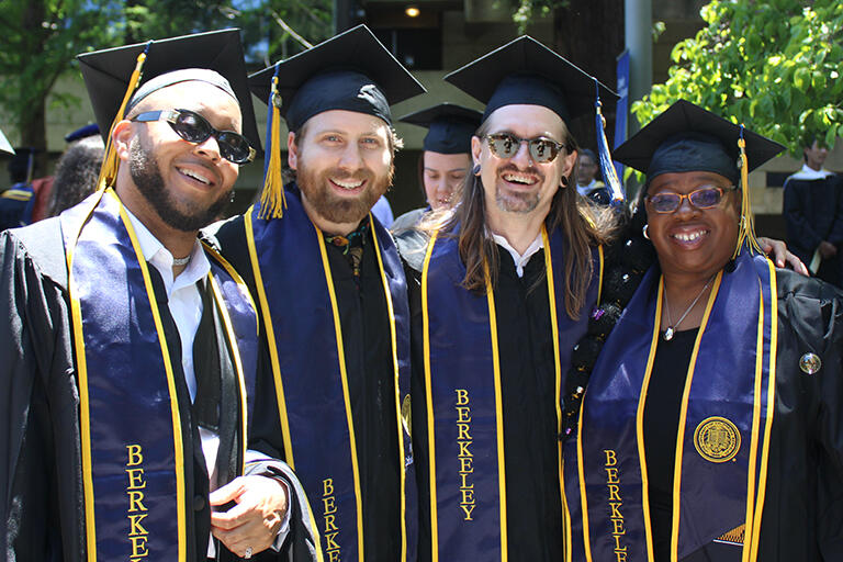 four master's degree students wearing their regalia before the ceremony standing next to each other smiling at the camera