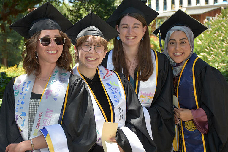 four master's degree students in their regalia lined up before the ceremony standing next to each other smiling at the camera