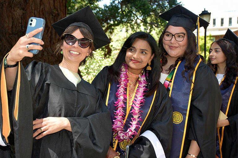 three master's degree students wearing their regalia lined up with the person in front taking a selfie of them