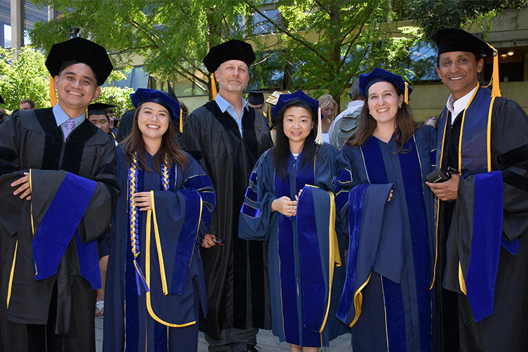 six doctoral candidates before the ceremony wearing their regalia standing next to each other holding their hoods and smiling at the camera