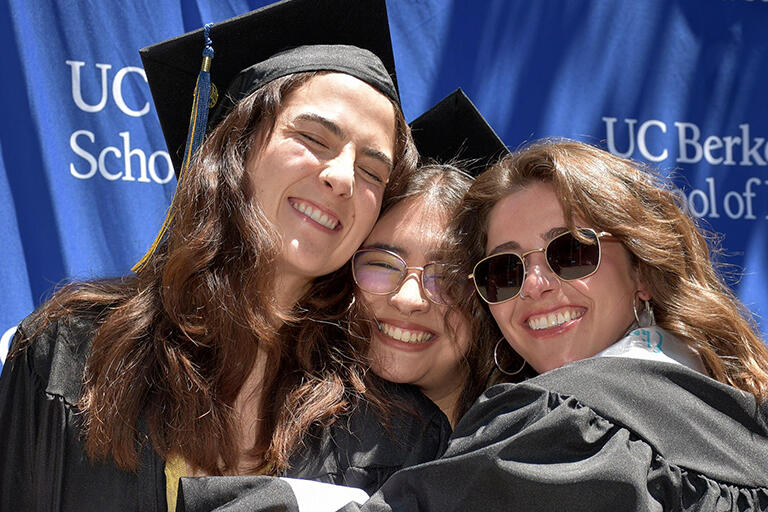 three graduates in front of the u c berkeley school of education backdrop hugging and smiling at the camera
