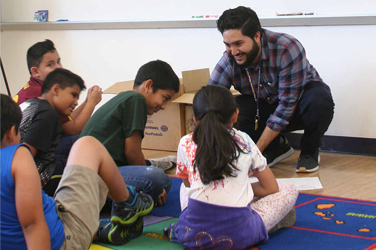 teacher smiling and squatting in front of students who are sitting on a colorful rug in a classroom